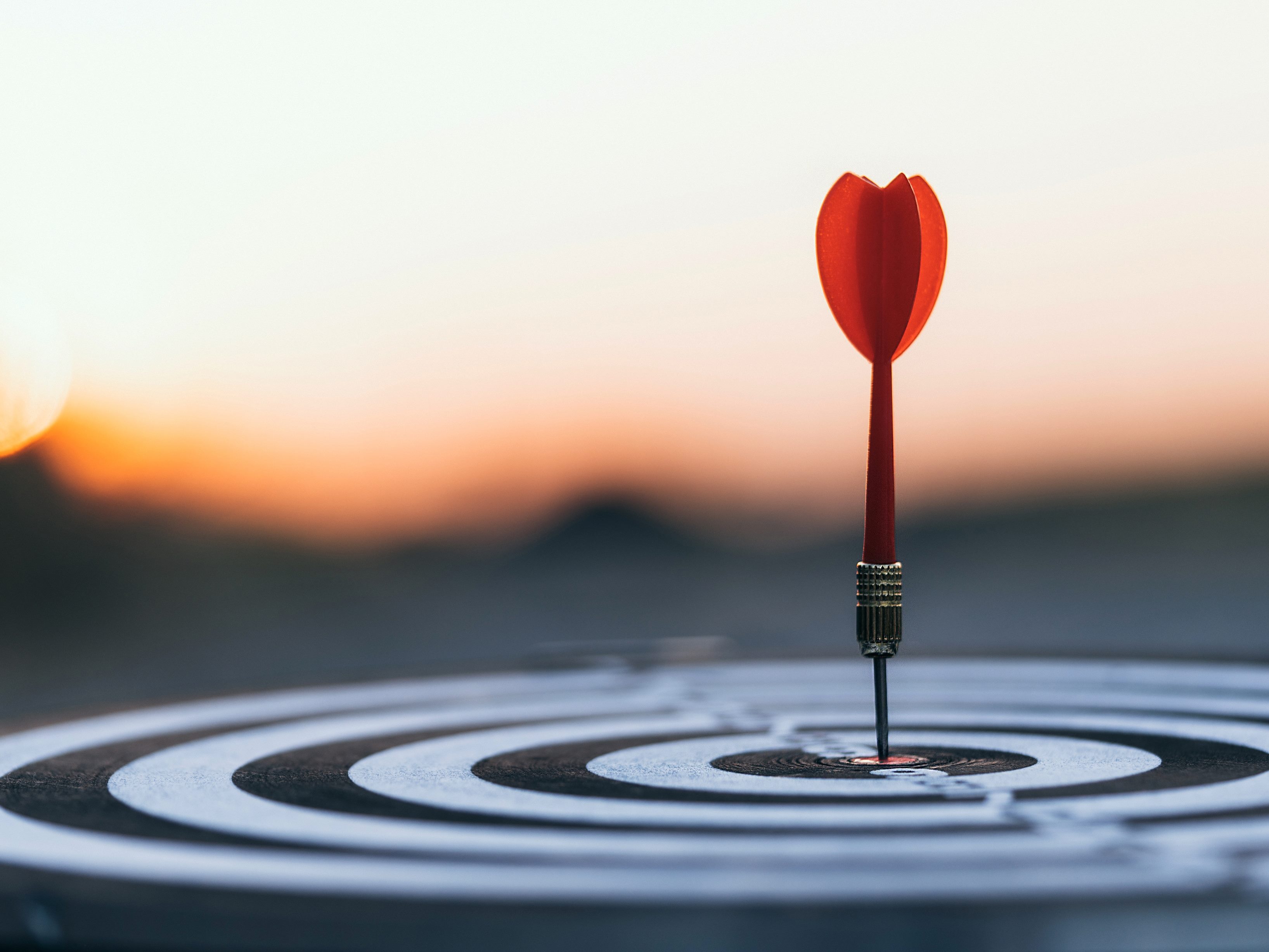 Close-up of red dart hitting the center target of a dartboard with alternating black and white rings, with a sunset blurred in the background. 