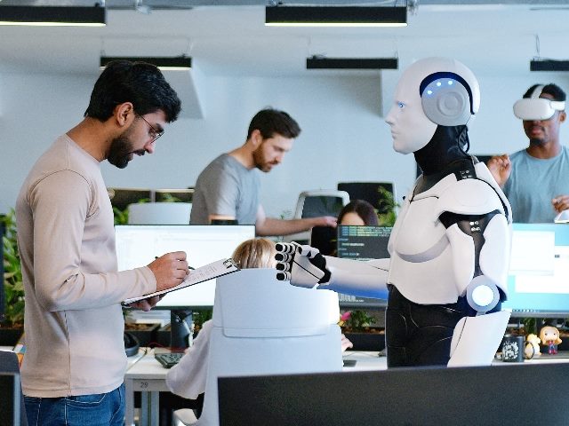 Man talking with robot in office, as two other humans work in the background, one to the right wearing an immersive device.