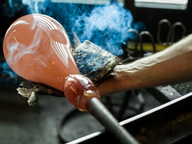 A glassblower shapes a piece of ocher glass using damp newsprint to shape the piece on the end of the punty, suggesting the power of AI martech tools.