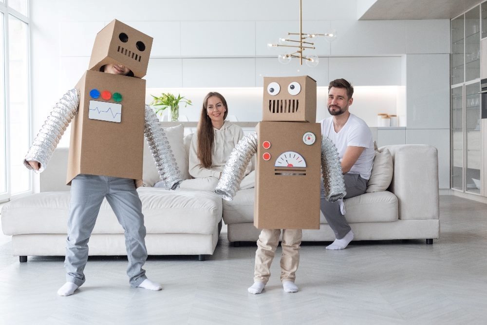 A family at home with the parents sitting on the couch and two children dressed in homemade robot costumes made from boxes and found objects in piece about generative AI tools.