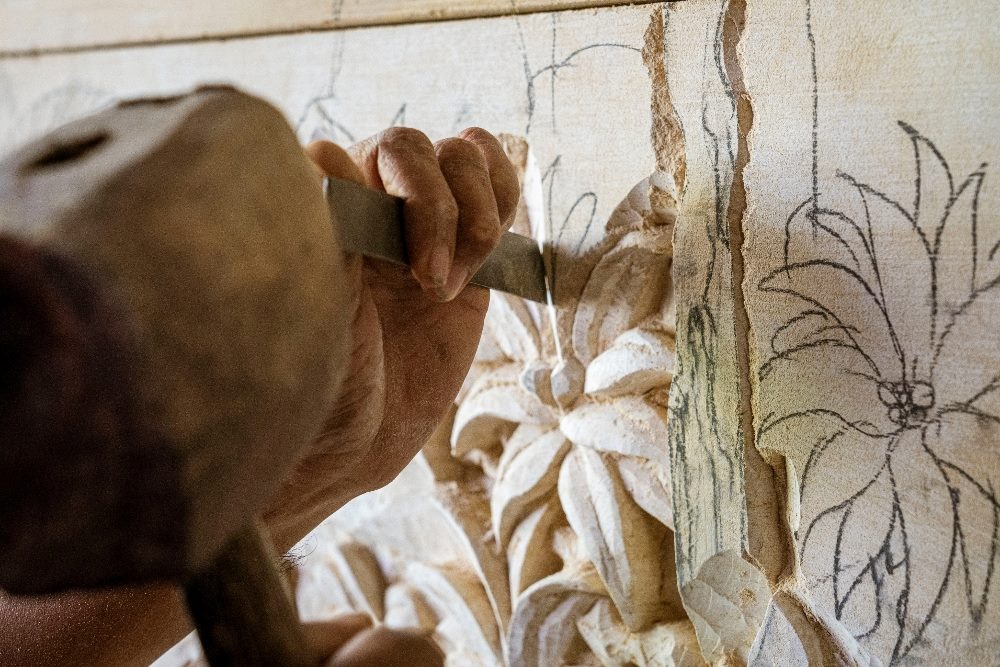 A close-up view of a skilled craftsman meticulously chiseling detailed floral patterns onto a wood panel, illustrating the concept of precise and deliberate craftsmanship akin to prompt engineering in AI.