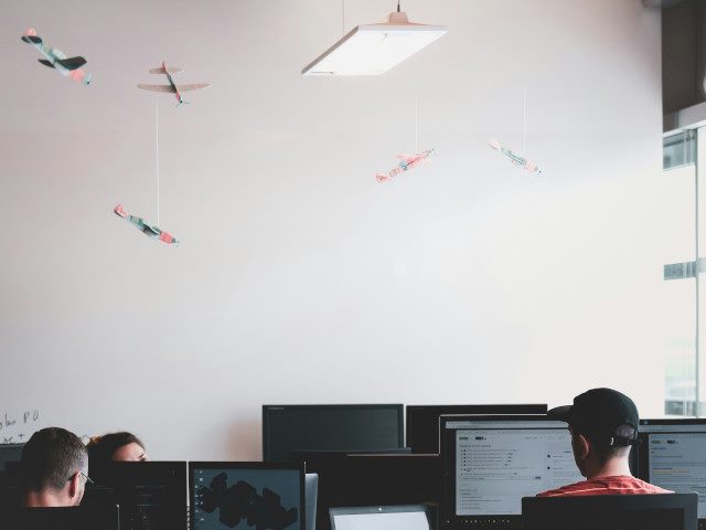 Employees work on computers at desks in an office with paper airplanes hanging from the ceiling.