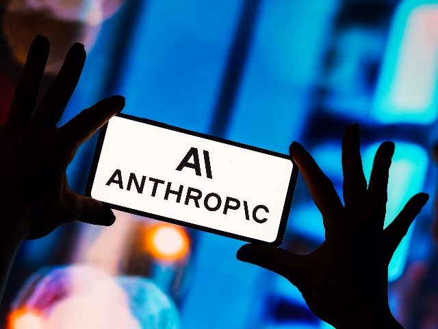 The Anthropic logo is displayed on a smartphone screen held in two hands amid bright blue lighting, in piece reporting on Anthropic's new deal with Amazon.