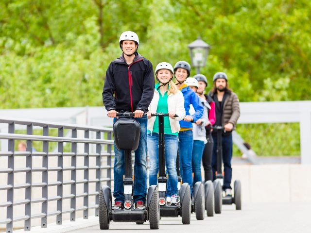 A group of six tourists ride Segways on a tour with blurred large trees in the background.
