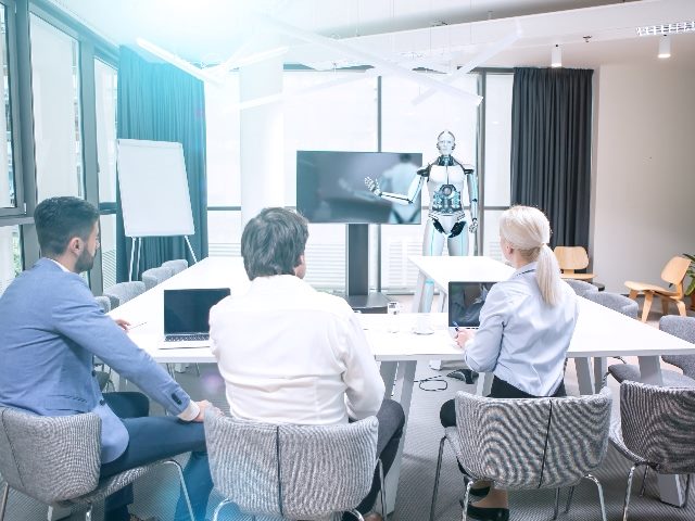 A humanoid robot leads business meeting while holding a pointer in front of a white board while three co-workers give him their attention while sitting at a table facing him, suggesting AI in CMO strategy is impacting the role. 
