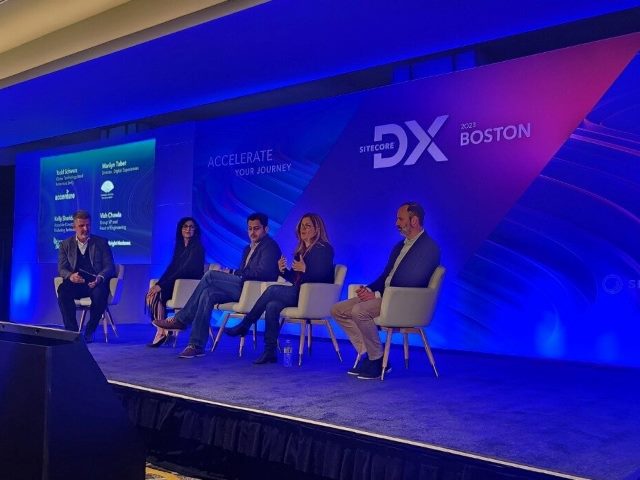 Sitecore DX Boston, people seated on a blue-lit stage with the words "Sitecore DX Boston 2023" on the back wall. 
