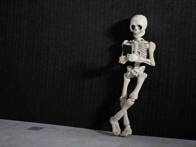 A skeleton leaning on a grungy wall with smartphone in his hand. 