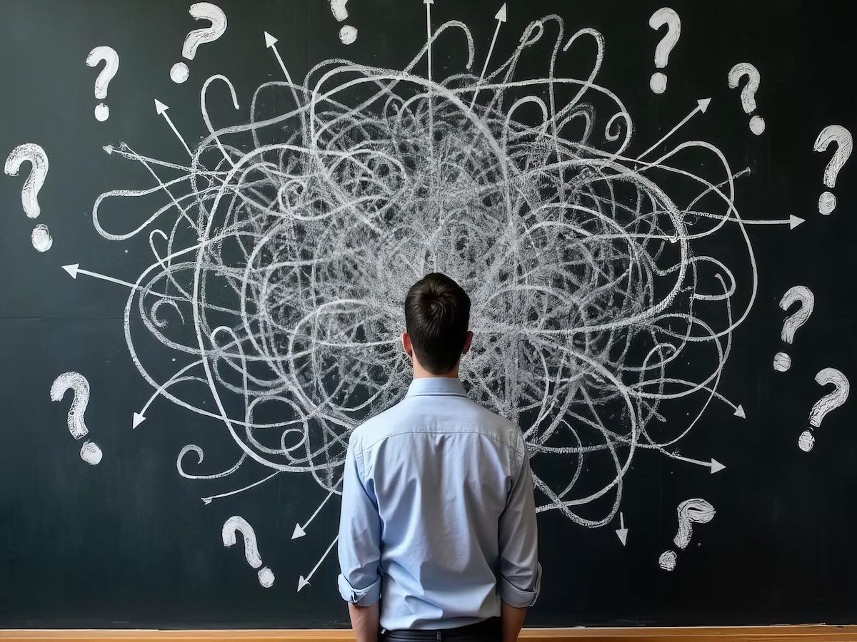 A person standing in front of a chalkboard filled with scribbles and question marks 