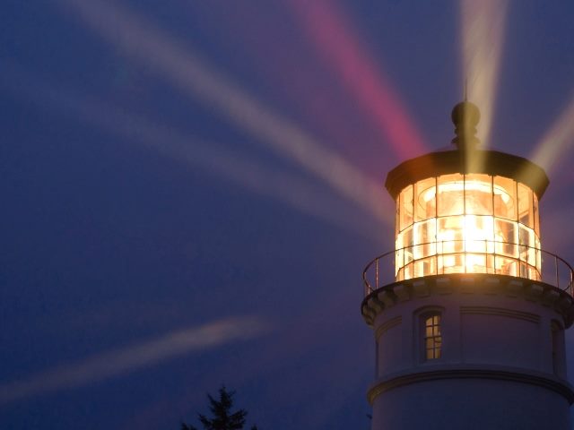 A light house beams golden  light out into the night during a rain storm with the outline of dark trees seen at the bottom on the horizon. 