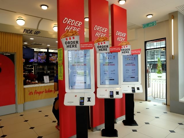 KFC self-service kiosks for ordering with order pickup area in background inside restaurant