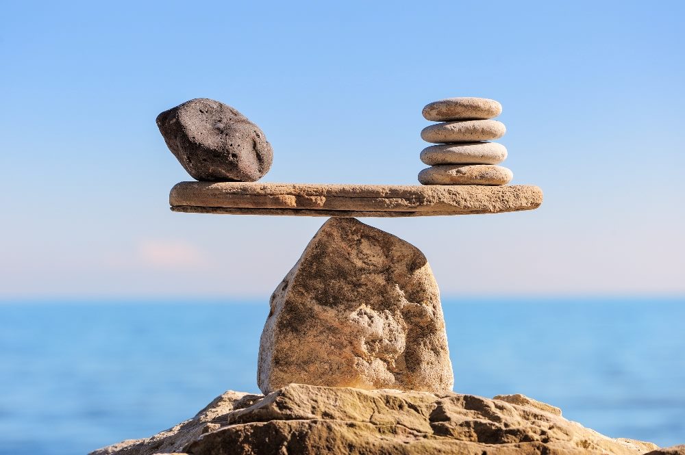 A flat rock balances a heavy single rock on the left and several flat disc shaped rocks on the right, resembling a balancing scale, in piece about AI and ethics in marketing.