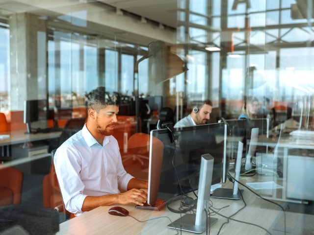 People working at a call center seen behind reflective glass and working at long tables on laptop computers in modern call center building.