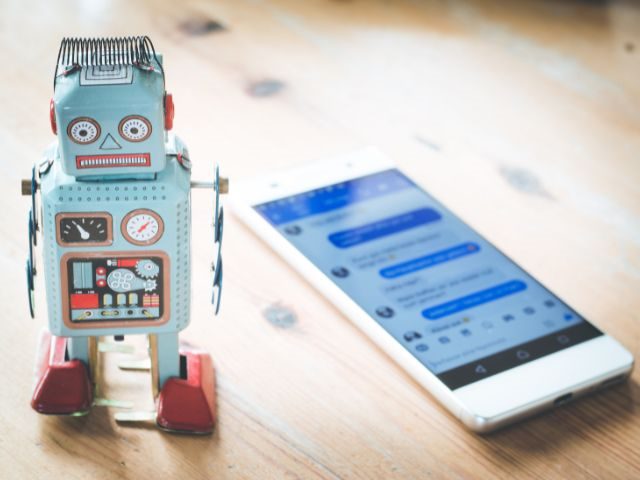 A small robot standing next to a phone with a customer service chatbot on screen