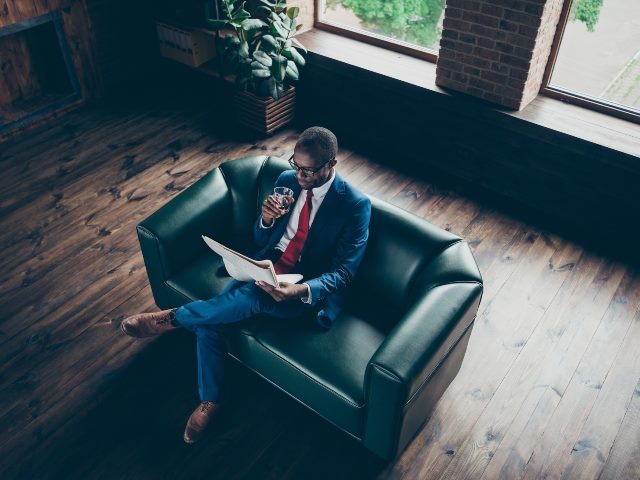Man sitting on a couch with a drink reading a newspaper.