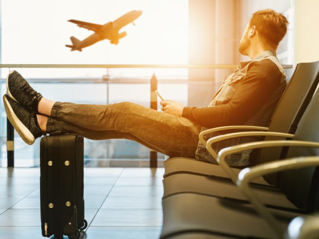 man sitting on a chair in an airport lounge with feet on luggage watching a plane take off