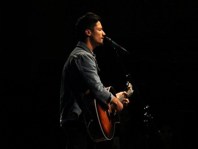 A singer-guitarist sings into a microphone on a stage.