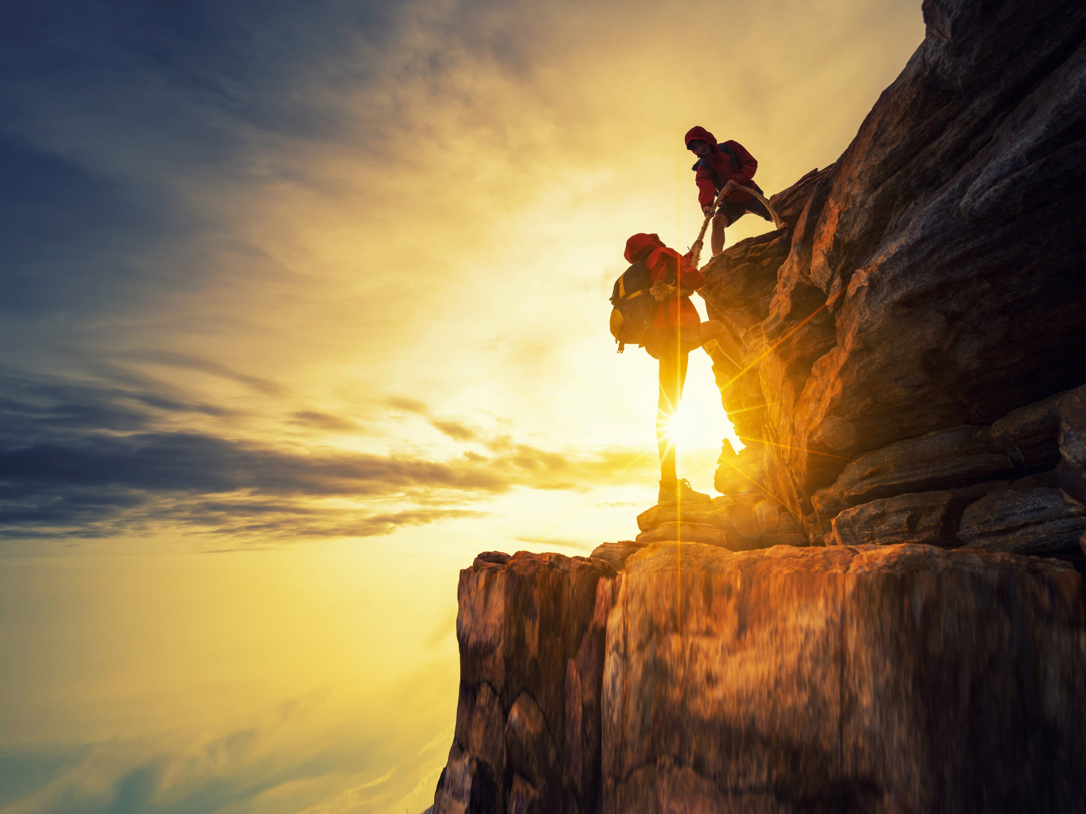 Couple hiking up a rock helping each other with silhouette in mountains with sunlight.