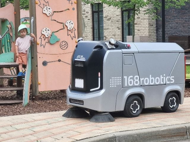 A child on a playground set looking at a small street sweeper van driving by.