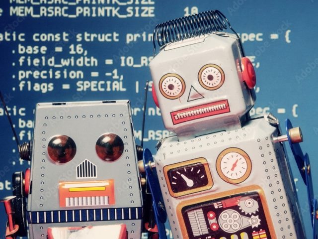 Two cute tiny toy silver robots with colorful accents in red, orange and yellow stand still as if in a selfie shot with computer coding, white on blue in the background.