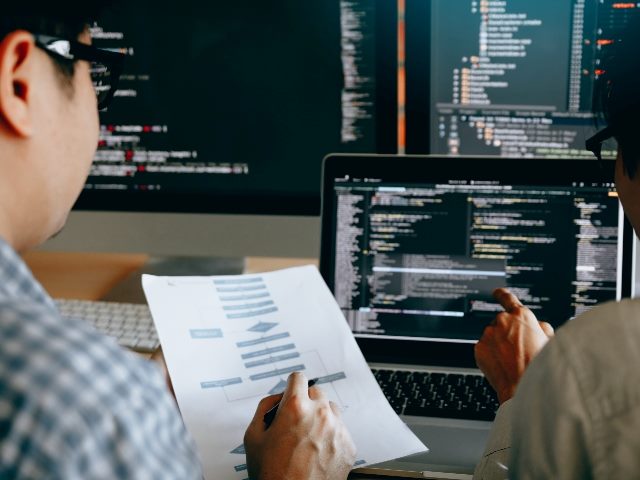 Two programmers/coders/developers, viewed from behind as they sit down at looking over paperwork and computer terminals, work on developing applications.