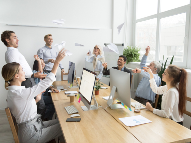 Multiracial corporate team of young and senior business people launching paper planes together in office, diverse motivated employees group laughing having fun at work