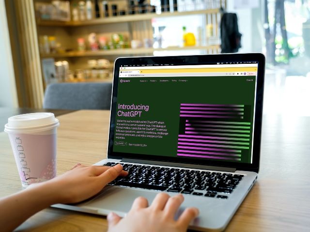 Person (you can only see their hands) works on a laptop with a coffee to the left. OpenAI home page for ChatGPT on the laptop screen. 