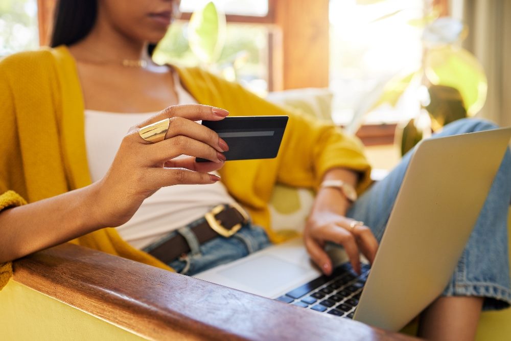A female customer wearing a gold sweater and white shirt and jeans holds a credit card while shopping on her laptop, suggesting the need for ethical AI principles for targeted customer communications.