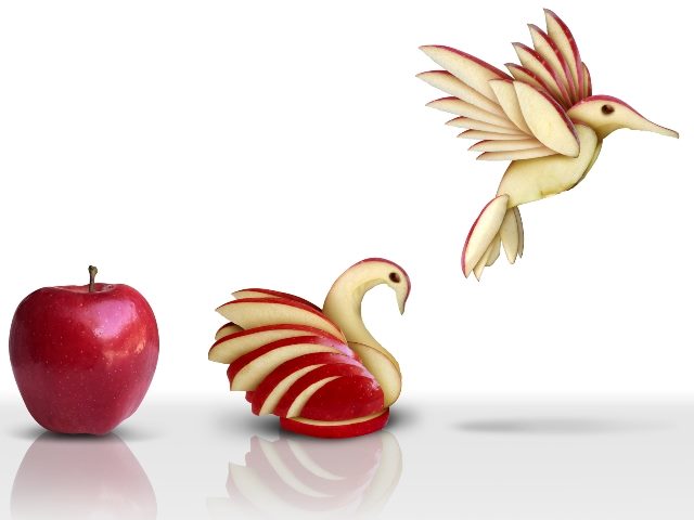 A red apples sits on a table on the left, followed by a swanlike bird carved from the apple that sits in the center of the table, and on the right another carved apple that is overtop the table that appears to be taking flight.