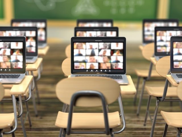 School desk and laptops with classroom app on the screen and blurred blackboard in the background. 