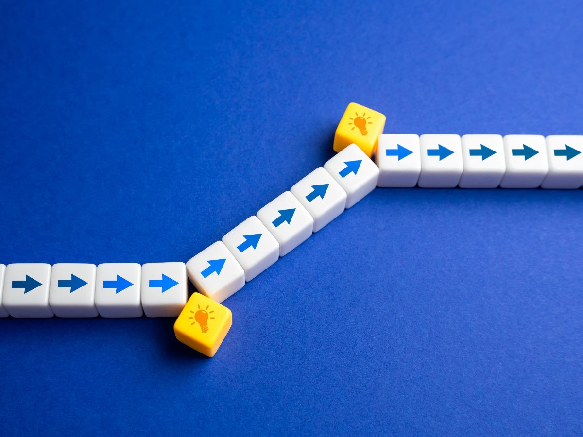 Conceptual image of white blocks with blue arrows in a straight line being redirected by yellow blocks with lightbulb icons, symbolizing innovation or a shift in direction.