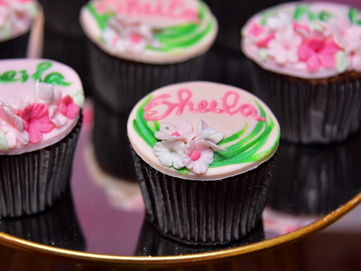 Close-up of decorated chocolate cupcakes displayed on a mirrored serving tray. The cupcakes feature pale pink icing with green accents, delicate flower decorations and piped names in pink frosting, creating a festive and personalized dessert presentation.