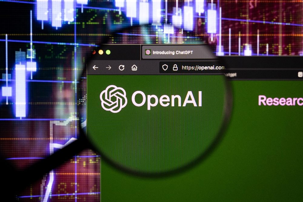OpenAI chatbot company logo on a website with blurry stock market developments in the background, seen on a computer screen through a magnifying glass in piece about technology predictions for 2024.
