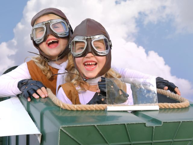 Two young girls wearing aviator cap-and-goggle sets sit in a toy "airplane" as pilot and copilot, against a backdrop of a sun-and-clouds sky. 