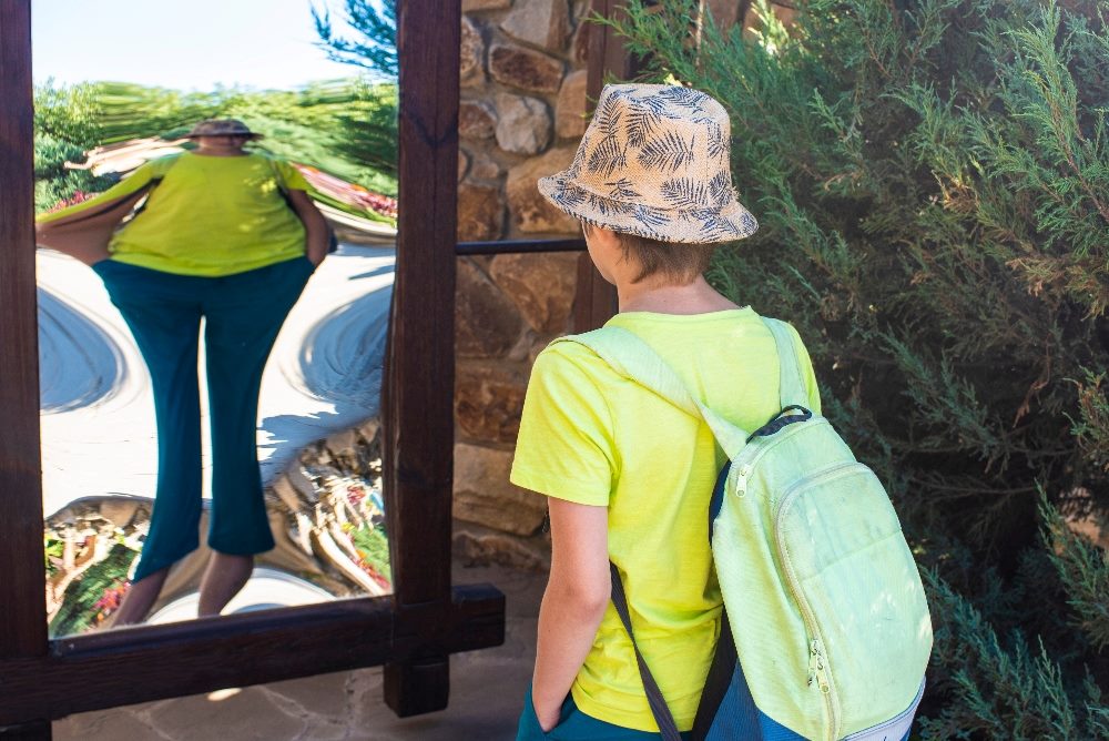 A young person with a light green backpack and a patterned hat is seen from behind, observing the distorted reflection in a funhouse mirror. The mirror dramatically alters the person's shape against a backdrop of lush greenery, illustrating the deceptive nature of AI content.