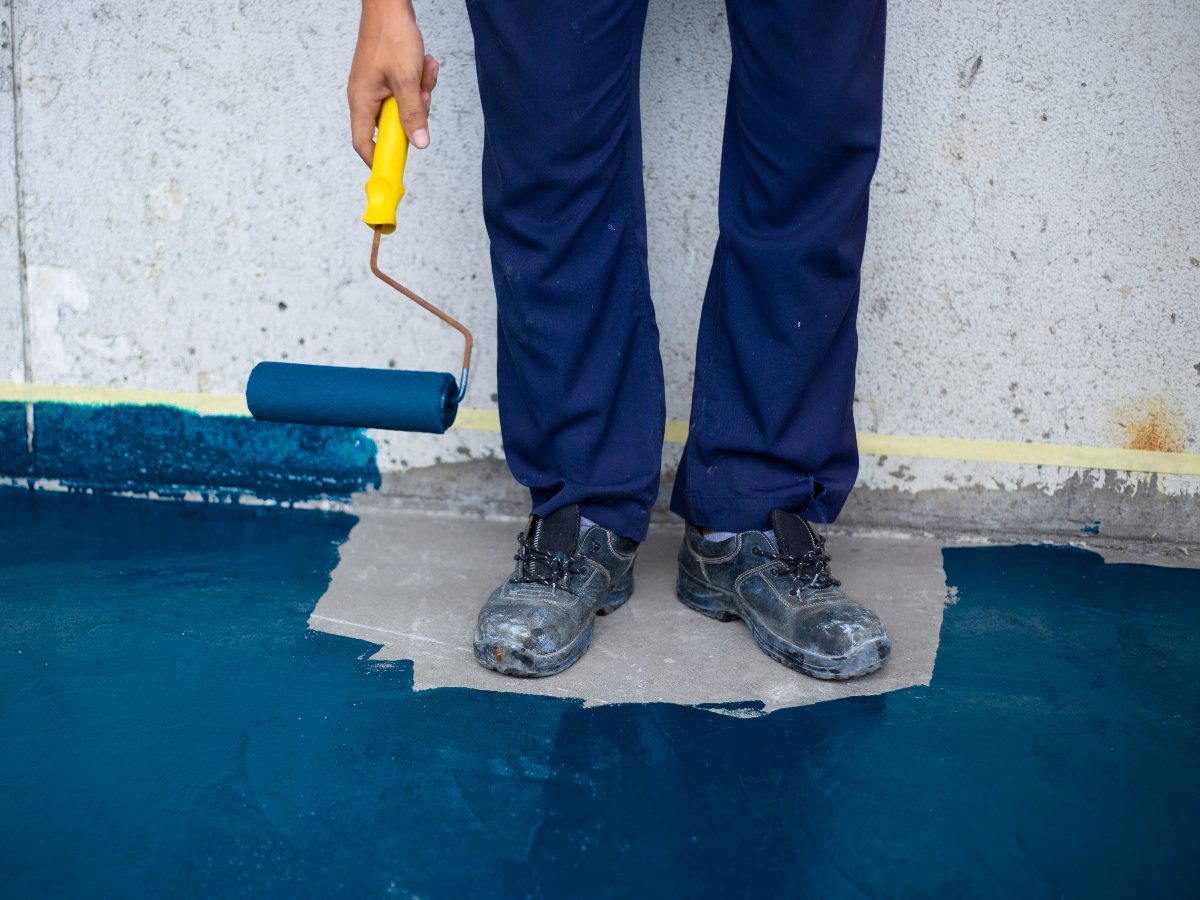 Person in work boots and blue pants painting a concrete floor with a blue paint roller.