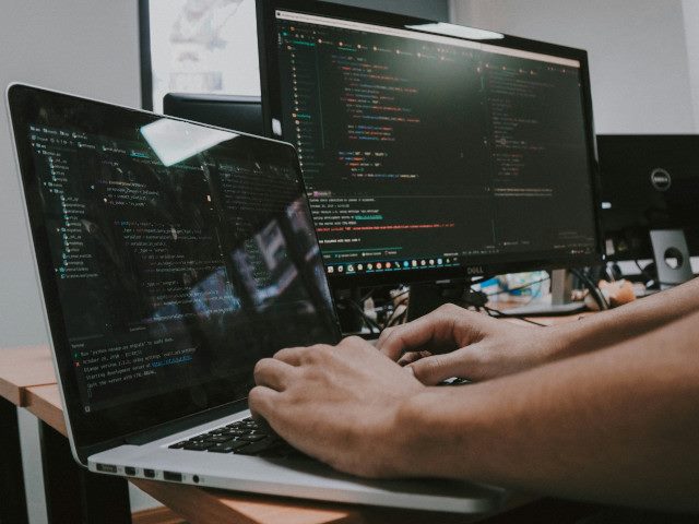 An employee codes software on a laptop computer and an external monitor on a desk in an office.