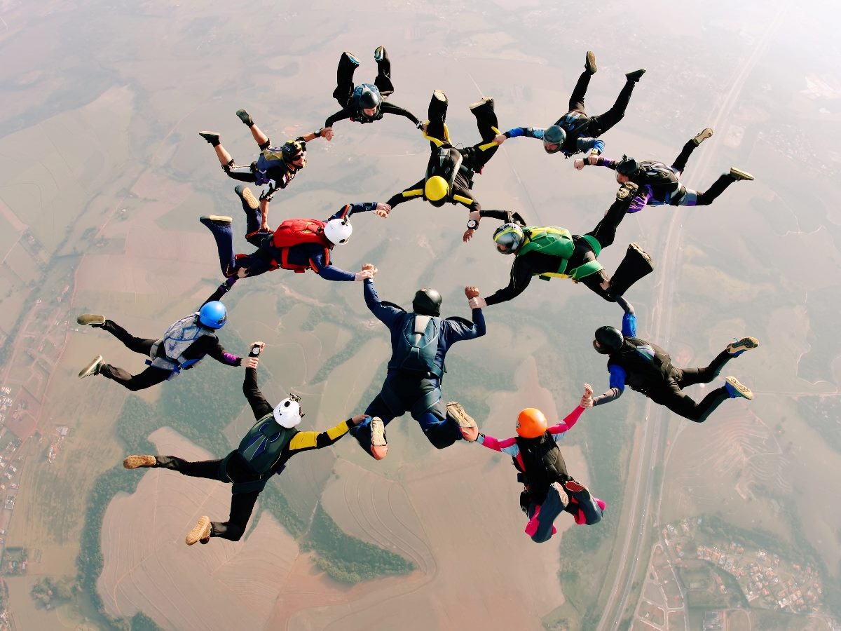 A group of skydivers in midair form a circle by holding hands while free-falling above farmland, illustrating coordination, trust, and shared risk.