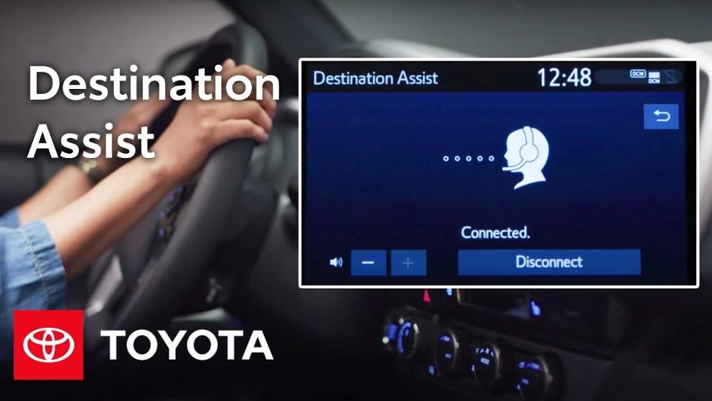 An image showcasing Toyota's Destination Assist feature. The image shows a close-up of a vehicle's dashboard screen displaying the Destination Assist interface, which indicates a connected status with an icon of a headset-wearing assistant and the options to increase or decrease volume and disconnect. The screen shows the time as 12:48. In the background, a person's hands are visible on the steering wheel, emphasizing the in-car use of the feature. The Toyota logo is prominently displayed in the bottom left corner, with "Destination Assist" written in large white text above it in piece about AI in CX.