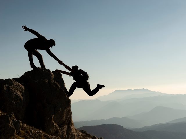 Silhouette image of one person on top of a mountain, grabbing the hand of another climber and lifting them up to the top. 