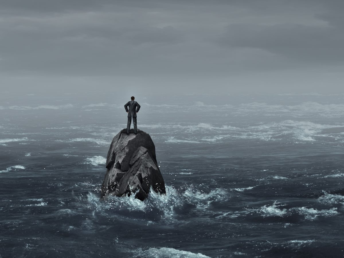 A lone businessman in a suit stands atop a jagged rock surrounded by rough, stormy ocean waves, gazing into the distance under a cloudy, overcast sky.
