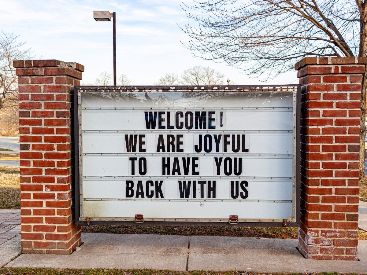 Outdoor marquee sign between two brick pillars reads, “WELCOME! WE ARE JOYFUL TO HAVE YOU BACK WITH US.”