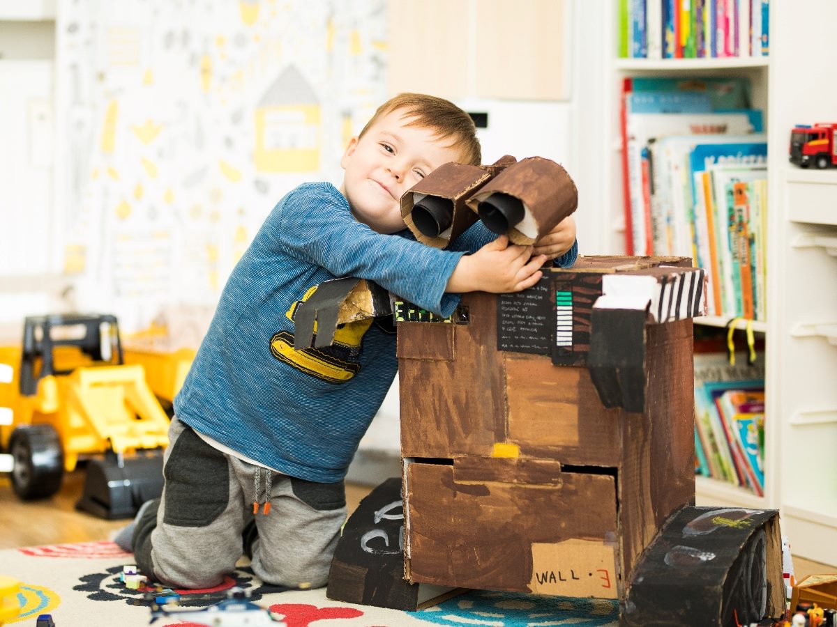 Child hugging a homemade cardboard robot toy resembling WALL-E in a playroom, with bookshelves and toy vehicles in the background.