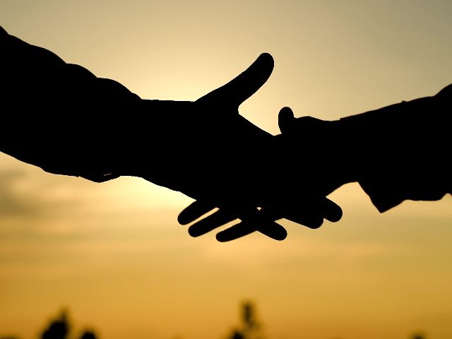 Silhouette of two people shaking hands with only the arms and hands visible against a gorgeous sunset on a body of water in the background in piece about AI in customer experience and trust.