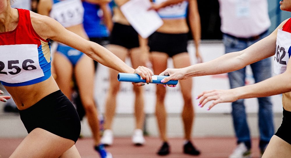 In a dynamic relay race scene, two female athletes in a stadium are focused on the precise moment of passing a blue baton. The background shows competitors and spectators, highlighting the intense competition. This image metaphorically represents how generative AI is rapidly advancing, akin to a seamless baton handoff, underlining how Google is thriving in this fast-paced environment.