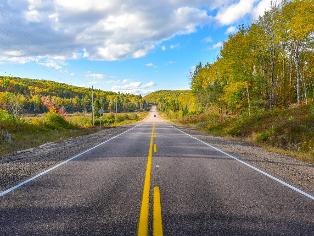 A long, empty rural road stretches into the distance under a partly cloudy sky, flanked by trees with early autumn foliage in shades of green, yellow and orange.