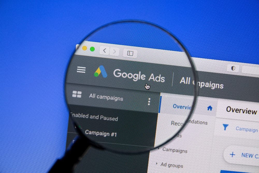 Google Ads website on a computer screen with a magnifying glass over it against a blue background in piece about Google Gemini AI model integration with Google Ads.