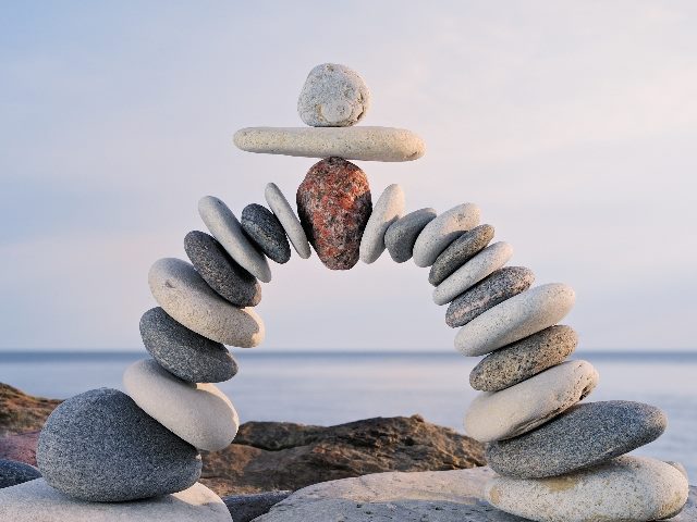 Smooth gray and white stones form an arch with a pink stone embedded in the middle on a rocky beach with water and the horizon in the background, suggesting need for balance in the ethics of AI in business.