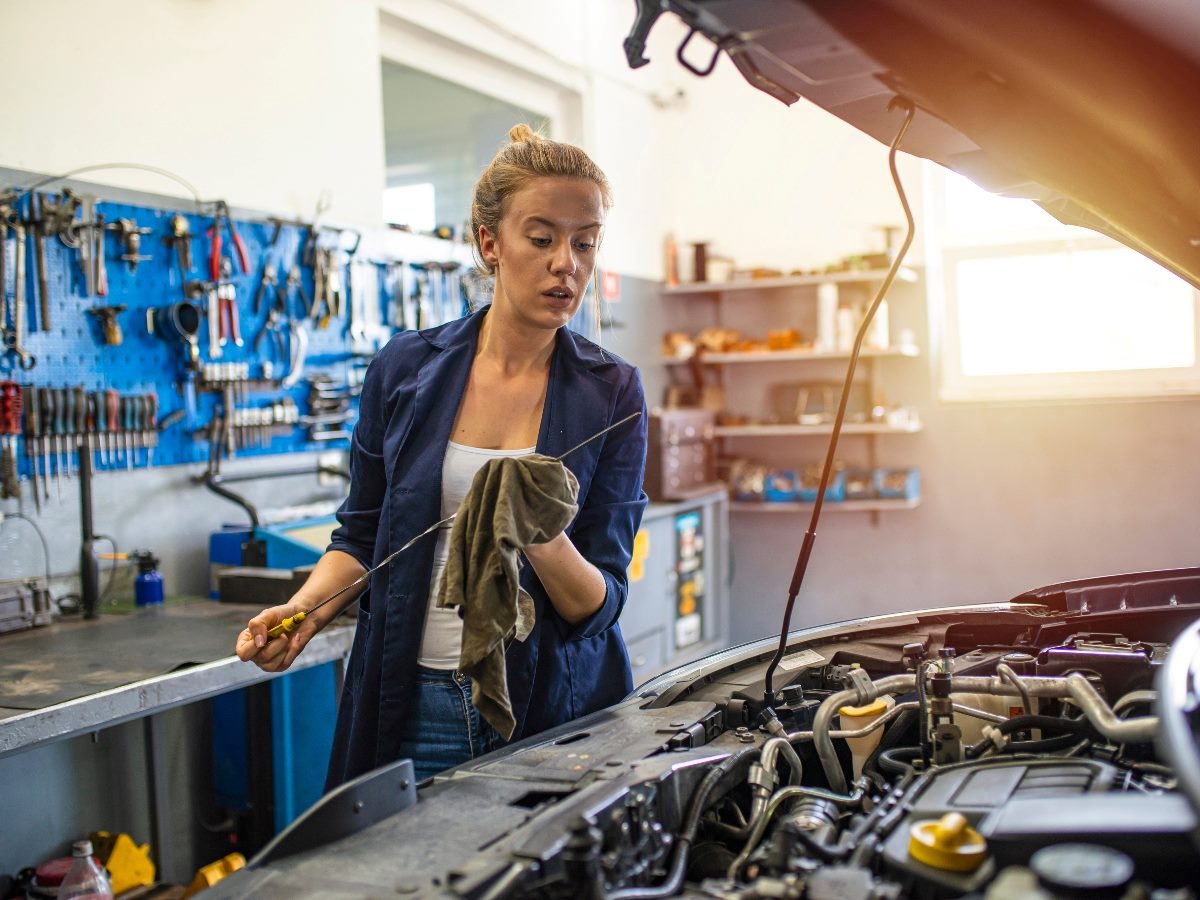Woman holding an oil dipstick looking under the hood of a car.