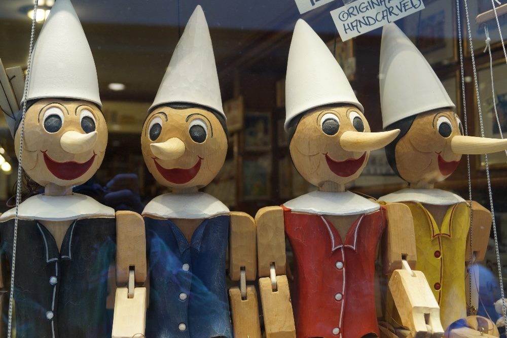 Four wooden Pinocchio puppets, two wearing blue jackets, one wearing a red jacket and one wearing a yellow jacket hang from strings in piece about generative AI platforms and the truth.