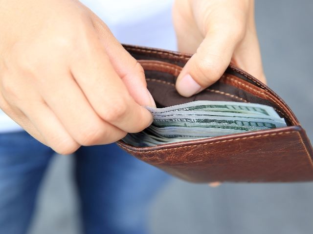 A man is opening his brown wallet to take money out, he's wearing blue jeans in a blurred background in story about customer experience technology purchases.